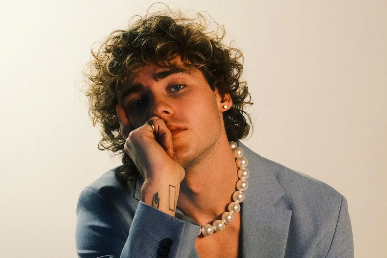 Portrait of musician Jack Gray with curly hair, wearing a pearl necklace and light blue suit jacket.